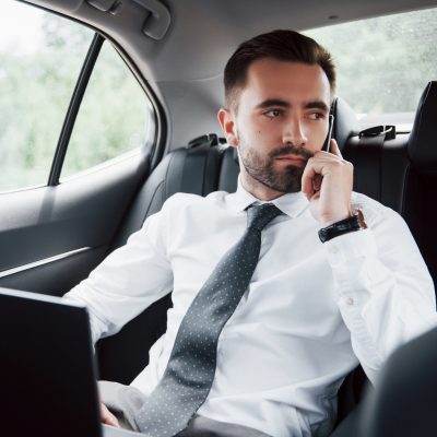 A young businessman working on laptop and talking on the phone while sitting in the car’s back. Works in motion, appreciates its time A young businessman working on laptop and talking on the phone while sitting in the car's back. Works in motion, appreciates its time.